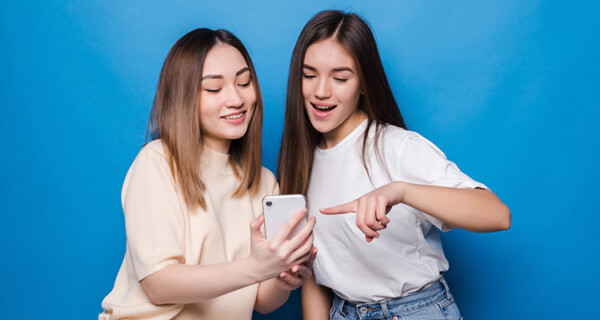 happy-two-young-girls-laughing-pointing-finger-screen-smartphone-while-taking-selfie-isolated-blue-wall